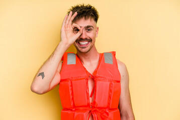 Young caucasian man wearing a life jacket isolated on yellow background excited keeping ok gesture...