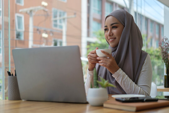 Thoughtful Muslim Business Woman Drinking Hot Coffee Sitting Front Of Laptop And Looking Out The Window, Thinking Work Plan.