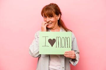 Fototapeta na wymiar Middle age caucasian woman holding I love mom placard isolated on pink background biting fingernails, nervous and very anxious.