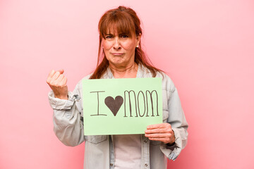 Fototapeta na wymiar Middle age caucasian woman holding I love mom placard isolated on pink background showing fist to camera, aggressive facial expression.