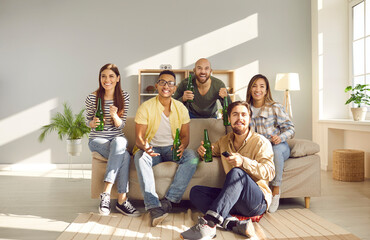Group of multiracial male and female friends watch sports competitions on TV at home. Emotional happy people with bottles of beer in their hands sitting on sofa and cheering for their favorite team.