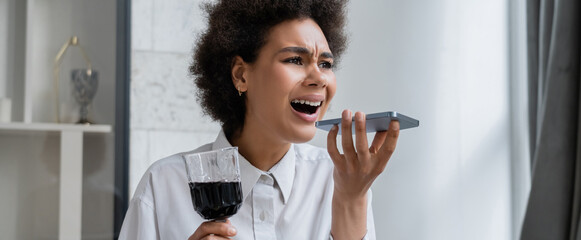 drunk and sad african american woman holding glass of red wine while recording voice message on smartphone, banner.