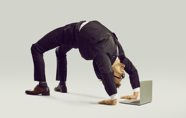 Happy energetic businessman practises yoga positions while working on laptop computer. Funny fit flexible young man in suit and glasses doing bridge pose on studio floor while using modern notebook PC - obrazy, fototapety, plakaty