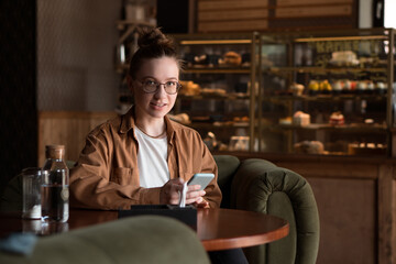 Young smiling woman sitting in cafe drinking coffe and using smartphone