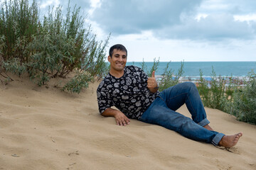 Fototapeta na wymiar Man sitting on the beach. A young, happy guy in jeans sits on a sandy beach near the sea. He looks into the camera and smiles happily.