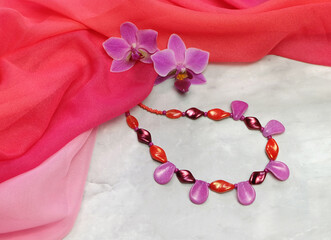 Czech glass beads with orchid flowers on a background of pink chiffon drapery, composition with handmade women's jewelry, with space for an inscription, selective focus, horizontal orientation.