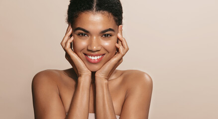 Smiling african american woman, applies daily care, nourishing facial mask on her skin and looking happy, standing over brown background