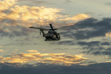 Poster Lage hoek opname van een militaire helikopter tijdens een vlucht in de bewolkte hemel bij daglicht tijdens zonsondergang © Grymen/Wirestock Creators