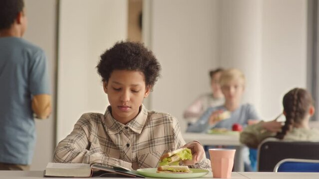 Waist Up Slowmo Of 11 Year Old African American Schoolgirl Reading Book While Having Lunch In School Canteen. Group Of Classmates Chatting At Table In Background