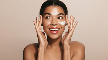 Happy smiling woman applies facial moisturizing cream, nourish her skin, stands over brown background - obrazy, fototapety, plakaty
