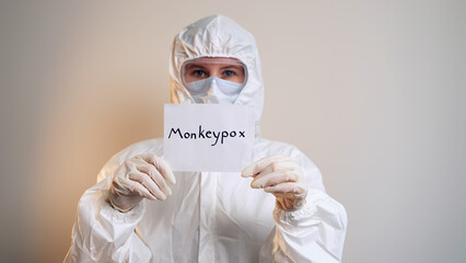 Isolated woman in protective suit, mask and goggles holding a sheet of paper with the inscription monkeypox close up. A new virus in medicine. Infectious disease. A new kind of pandemic. 