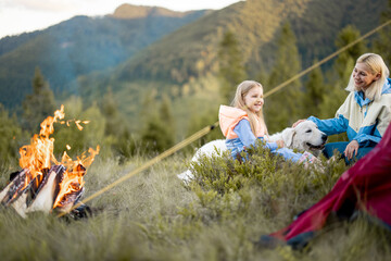 Young woman with her little girl and dog sit by the campfire, bonding together while travel on nature. Mother with daughter care pet at campsite