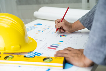 Asian engineer with architectural house plan project blueprint, graph and yellow helmet and...