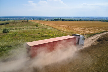 Fototapeta premium Aerial view of cargo truck driving on dirt road between agricultural wheat fields making lot of dust. Transportation of grain after being harvested by combine harvester during harvesting season
