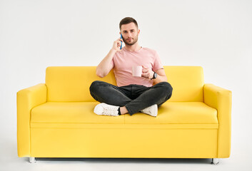 Fototapeta na wymiar Relaxed man talking on mobile phone and drinking coffee sitting on comfort bright sofa