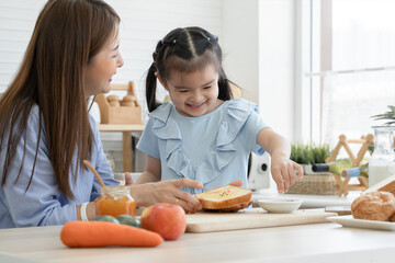 Happy Asian family, cute little daughter enjoy adding rainbow sugar flakes on sliced bread with jam with young mother while preparing breakfast together with fruits and fresh milk in kitchen at home