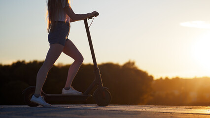 A beautiful slender young woman rides an electric scooter at sunset. Back view, copy space