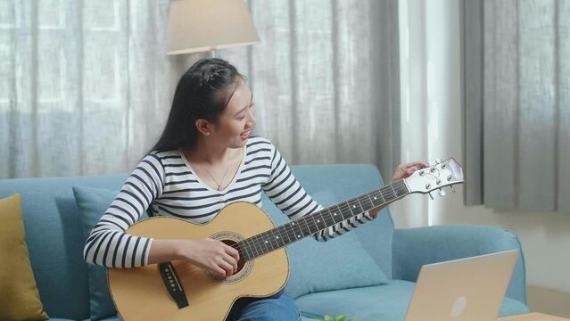 Asian Woman Learning Pulling Strings Of Guitar By Laptop At Home
