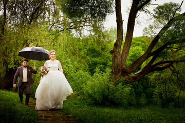 Wedding couple walks in green park with an umbrella in rain in a green summer day