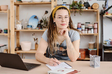 Young modern female in casualwear and eyeglasses sitting by workplace in studio and looking at...