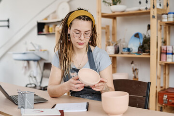 Young woman holding handmade earthenware over table while sitting in studio or workshop and...