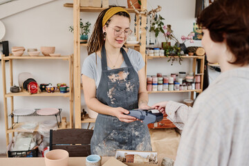 Young female owner of small earthenware shop with payment terminal selling handmade items to client paying for purchase by credit card