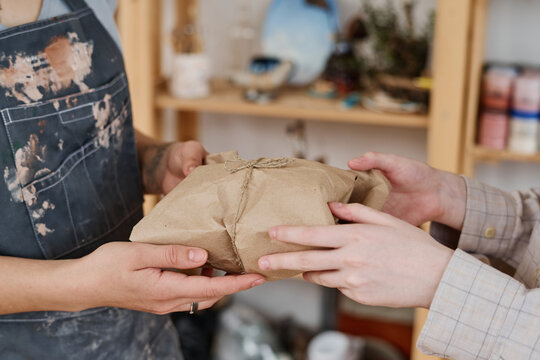 Hands of young craftswoman passing package wrapped into paper and tied by thread to female buyer in workshop or earthenware shop