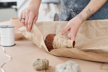 Hands of young female pottery master or shop assistant wrapping earthenware item ordered by client...