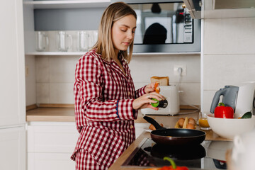 Young white woman wearing pajama standing by stove and cooking meal