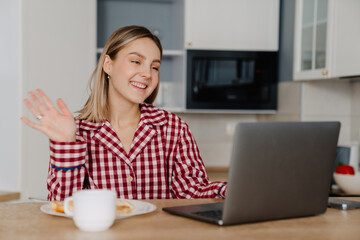 Young white happy woman having breakfast and using laptop in kitchen