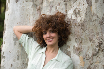Obraz na płótnie Canvas Portrait of attractive mature woman with curly brown hair and shirt, leaning against a tree trunk with one hand behind her head smiling happily. Concept happiness, hairstyle, curls, beauty.
