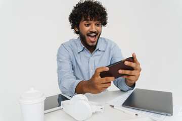 Young smiling indian man sitting at table playing phone