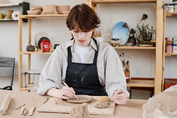 Brunette girl in black apron using wooden cutter for making ornament on clay plate while sitting by...