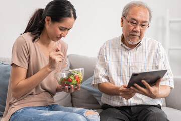 Obraz na płótnie Canvas Young Asian daughter serving and feeding food to senior father while he using and playing on digital tablet in the living room. family love and support to elderly people. 