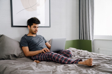 Young handsome indian man liying on bed working with laptop