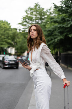 Trendy woman with mobile phone and suitcase on street in Paris.