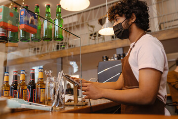 Young indian curly barista in face mask working with tablet