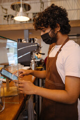 Young indian curly barista in face mask working with tablet