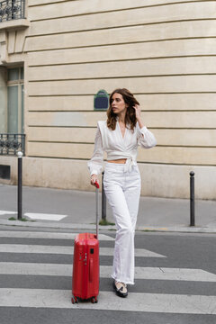 Trendy woman with suitcase walking on crosswalk on urban street in Paris.