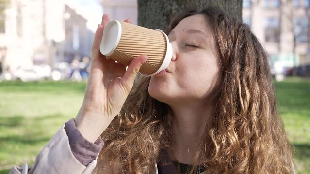 Young woman with curly loose hair drinks coffee in paper cup on blurred background. Female student enjoys lunch in city park before classes closeup