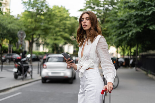 Stylish woman with cellphone and suitcase standing near road on street in Paris.
