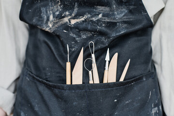 Part of craftsperson in black apron with handtools necessary for carving ornaments on handmade clay...
