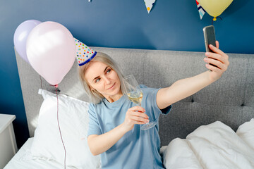 Happy emotional woman in pajama and party cap doing selfie shot on mobile phone at birthday morning while sitting on the bed with champagne and balloons in bedroom. Happy birthday concept