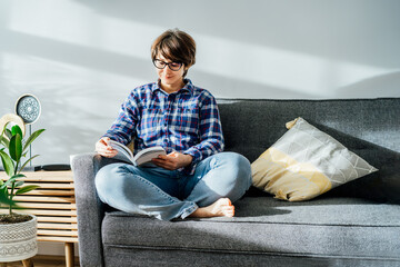 Woman in eye glasses sit on the sofa and relax while reading a book at home in cozy modern interior...