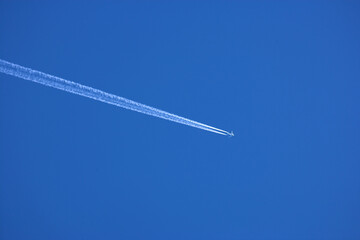 Airplane contrail against clear blue sky background with copyspace. View of a distant passenger jet plane flying on high altitude in blue sky leaving long white smoke trail behind. Air transportation