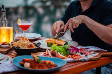 Man on luxury dinner served on table with fresh seafood, cold cuts and cocktail glass in luxury outdoor restaurant. Man hands cutting grilled salmon. Healthy food