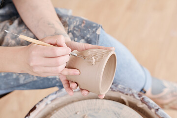 Hands of young female pitcher or ceramist with handtool carving patterns on sides of handmade clay cup while sitting by pottery wheel - Powered by Adobe