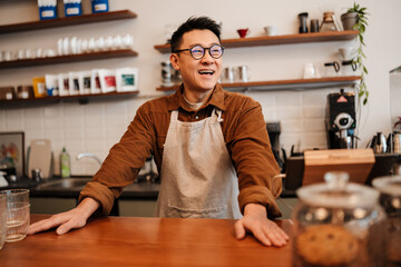 Adult happy asian man wearing apron standing at counter in cafe