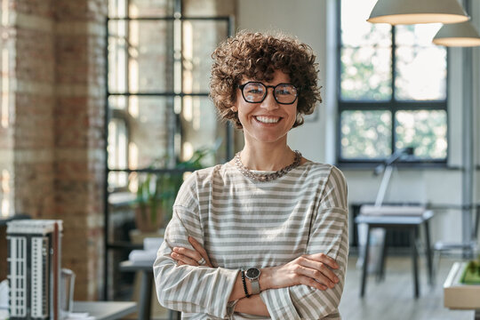 Portrait of young successful architect in eyeglasses standing with arms crossed at office and smiling at camera