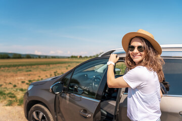 Happy cheerful young woman in hat and sunglasses traveling by car in the countryside on a summer day